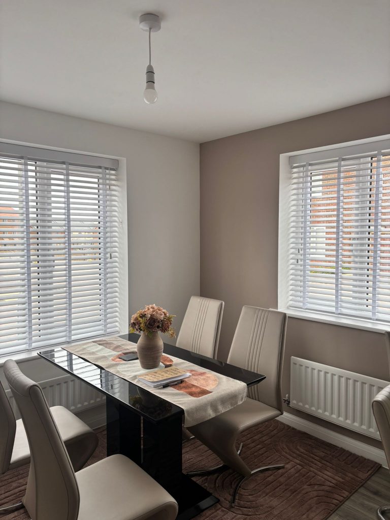 Modern dining room with six beige chairs, a glass table, floral centrepiece, and large windows with white blinds letting in natural light.