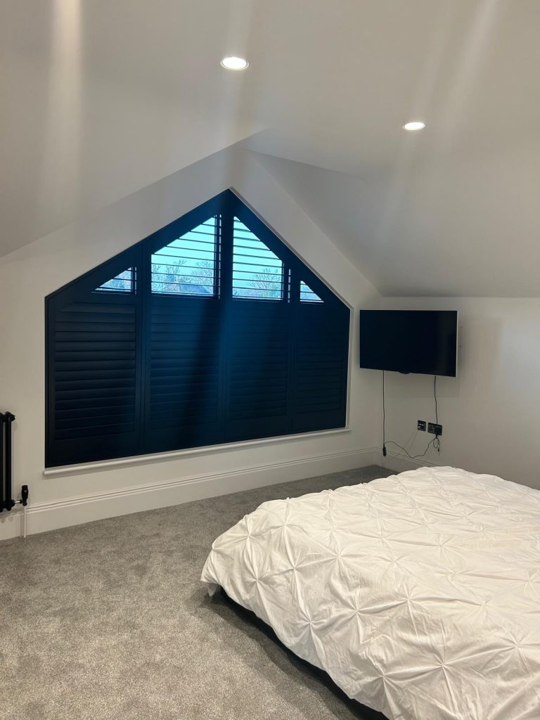 A modern bedroom with a large triangular window, black shutters, grey carpet, a white bed, and a wall-mounted telly.