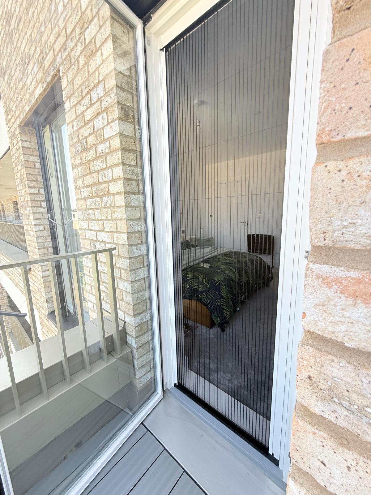 A balcony view shows a bedroom through a glass door with black insect screens, brick walls, and grey flooring.