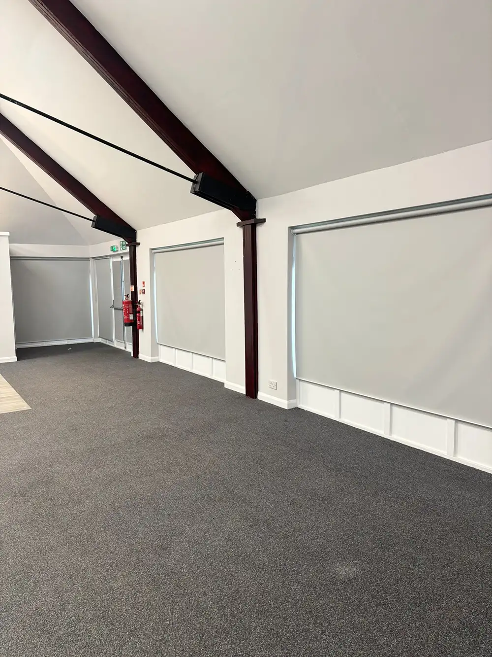 Spacious room with grey carpet, white walls, large roller blinds on windows, exposed beams, and fire extinguishers near the exit.
