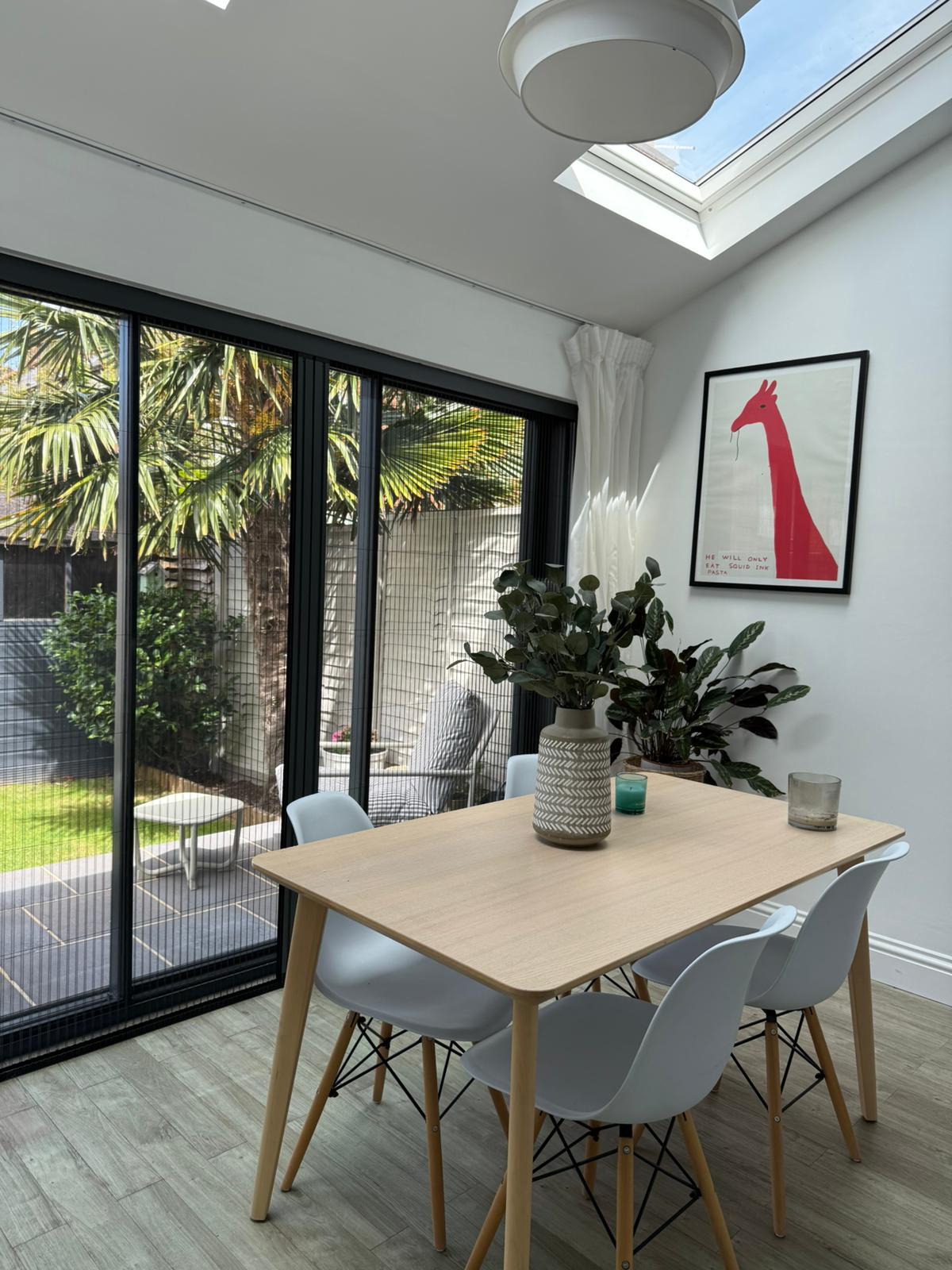 A modern dining area with a light wood table, white chairs, plants, and insect screens on large glass doors opening to a sunny garden.