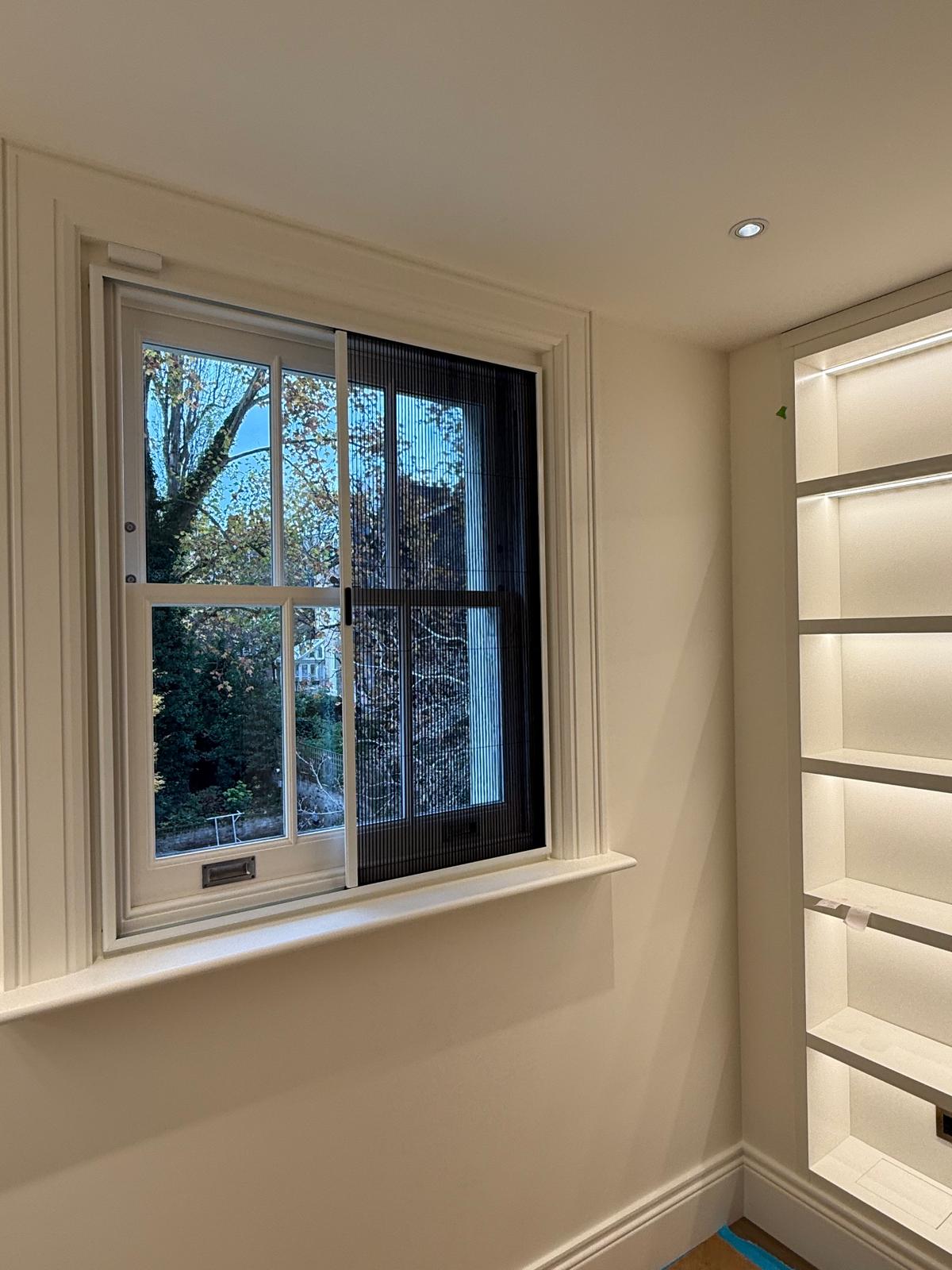 A window with insect screens, a white shelving unit on the right, and outdoor trees visible through the glass.