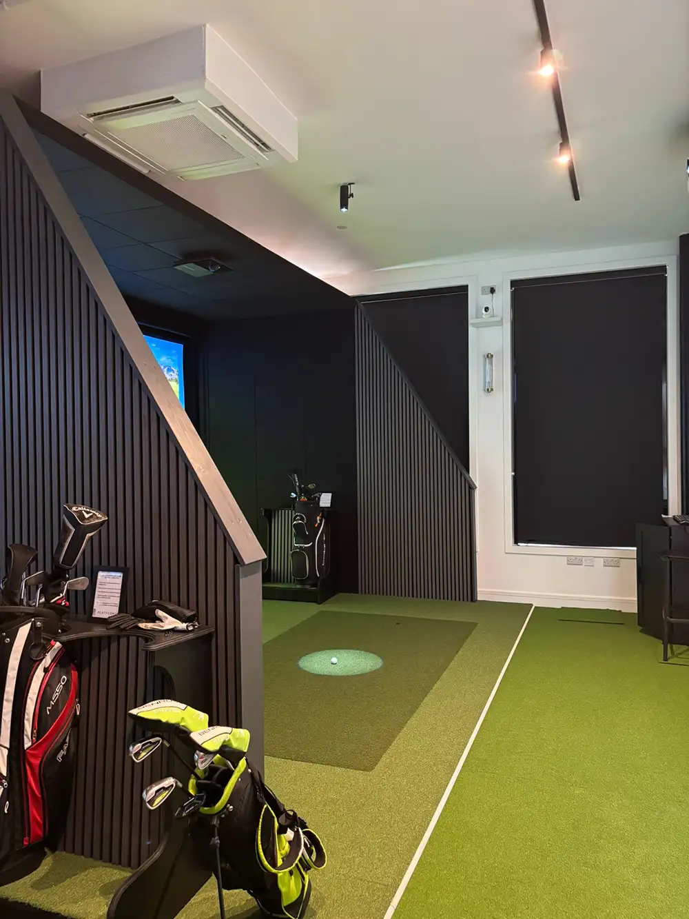 Indoor golf simulator room with golf bags, green turf, golf clubs, and Made to Measure Blackout Roller Blinds for light control.
