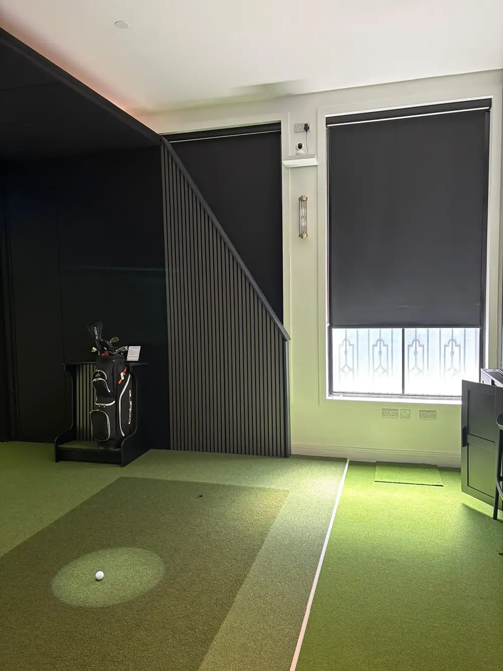 Indoor golf simulator room with green turf, golf gear, and two large windows fitted with Made to Measure Blackout Roller Blinds.