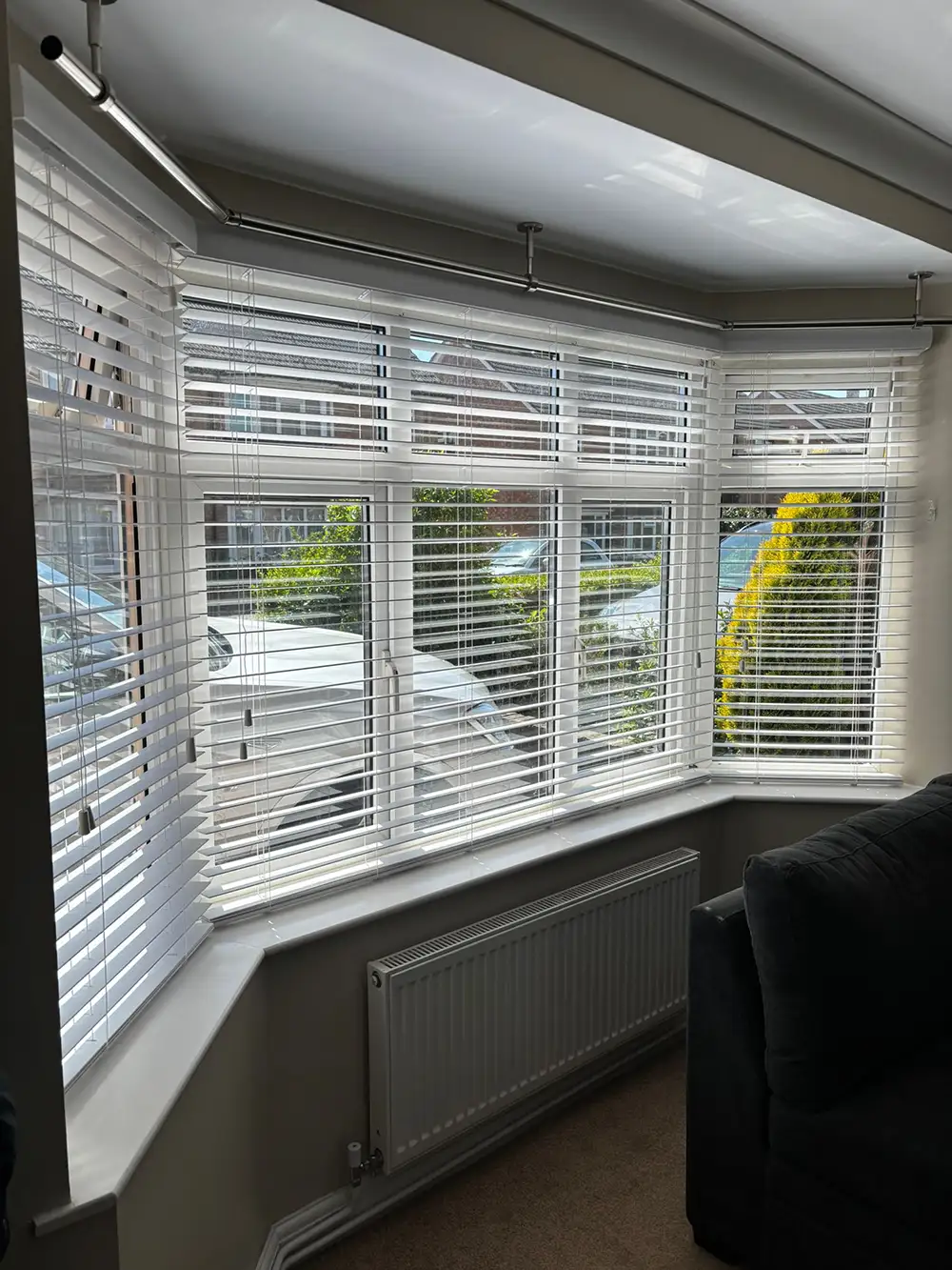 A bright bay window with white Venetian blinds, overlooking parked cars and greenery, plus a radiator and sofa inside.
