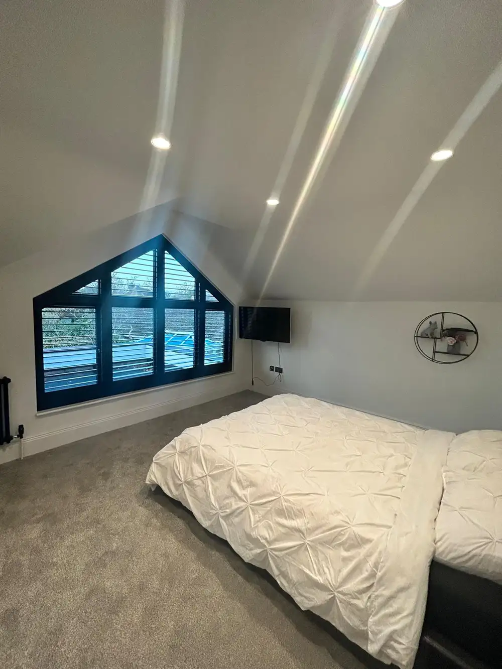 A modern loft bedroom with shaped shutters on large triangular windows, a bed with white duvet, TV, and a round wall mirror.