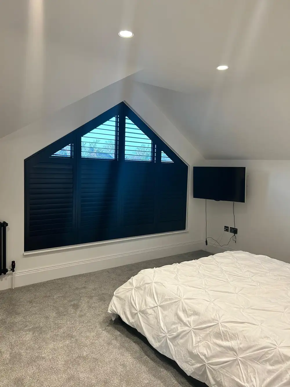 Modern bedroom with a sloped ceiling, large triangular window with shaped shutters, wall-mounted telly, and a white bedspread.